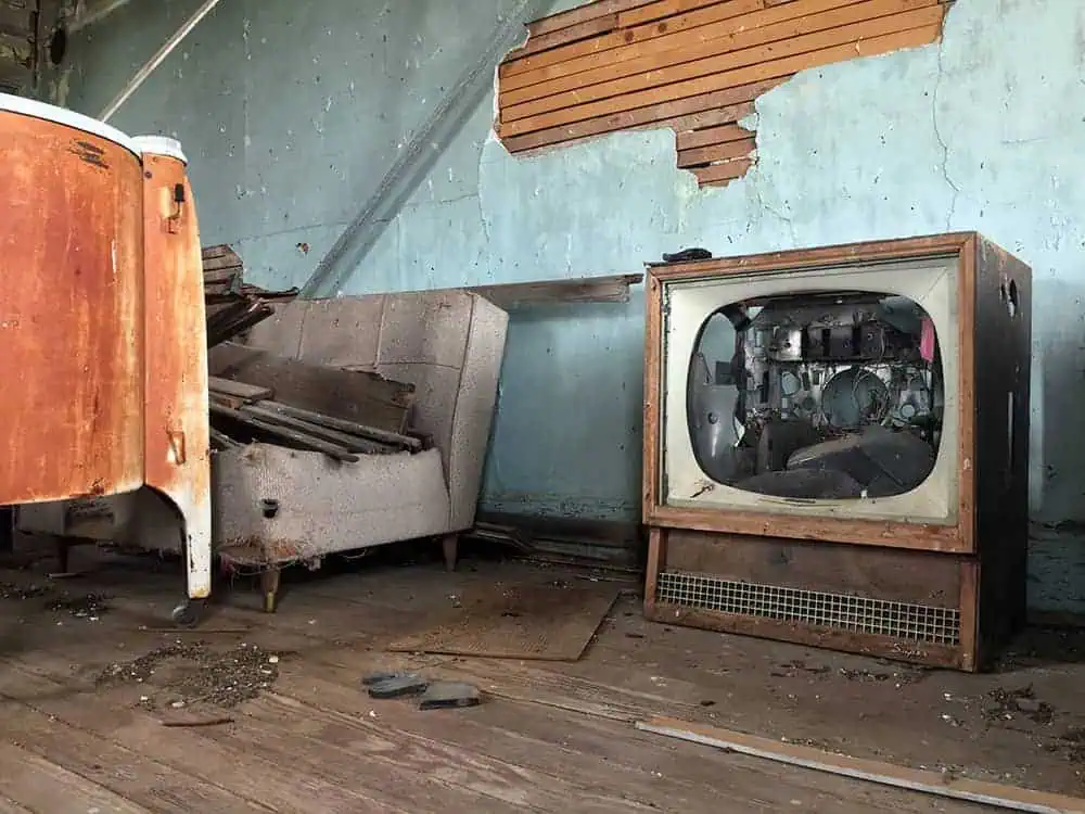 A decaying room with a broken vintage television, torn sofa, and peeling blue wall exposing wooden lath beneath damaged plaster.