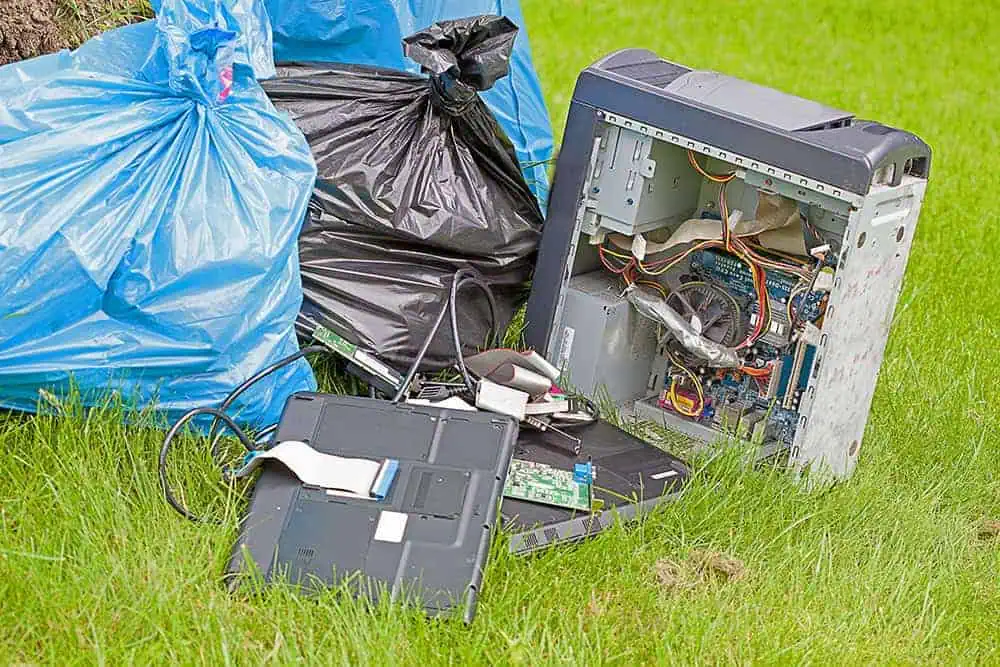 Discarded desktop computer and laptop with exposed parts beside garbage bags on green grass.