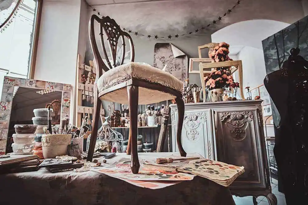 Vintage chair flipped upside down on a worktable in a cluttered studio with paint swatches, brushes, and ornate cabinets.