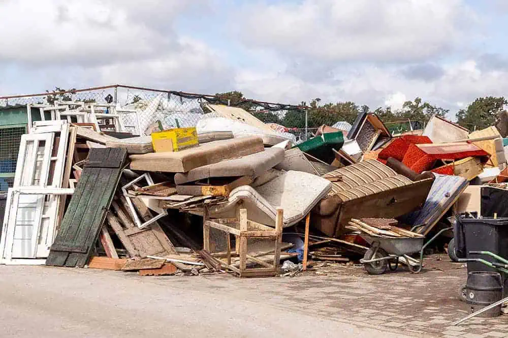 A large pile of discarded furniture, wood, and household items stacked at a junkyard under a cloudy sky.