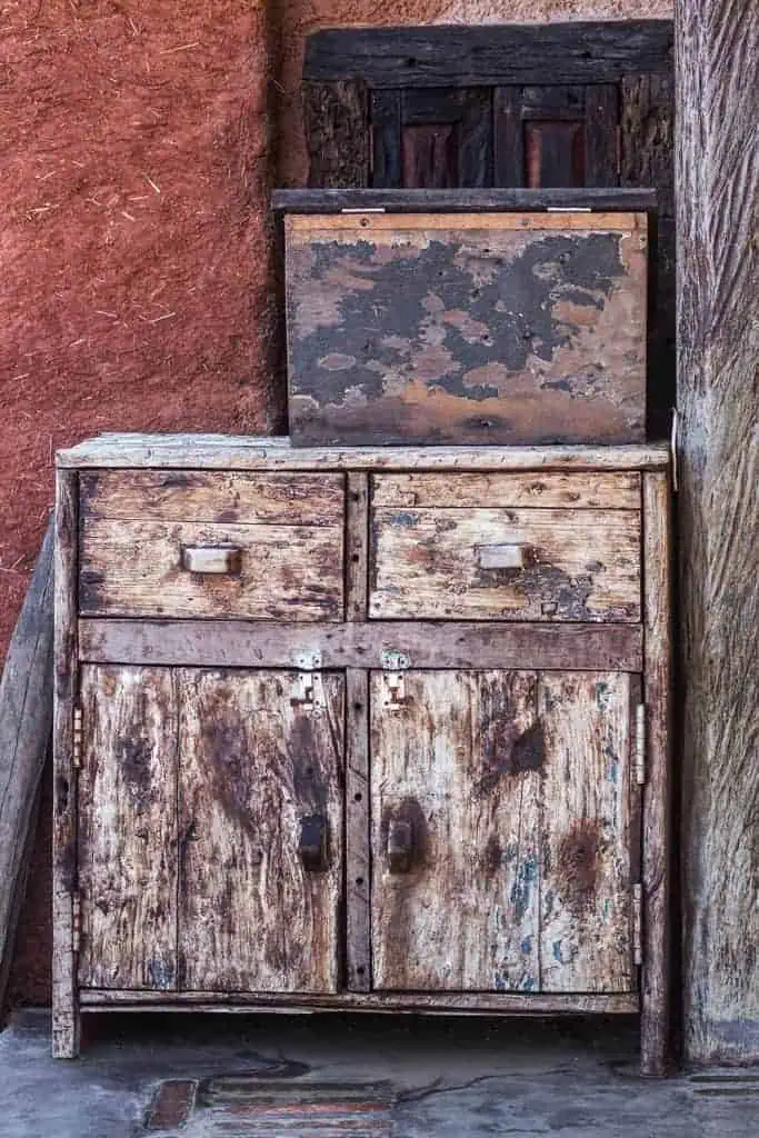 A weathered wooden cabinet with peeling paint and rustic textures against an earthy red wall.