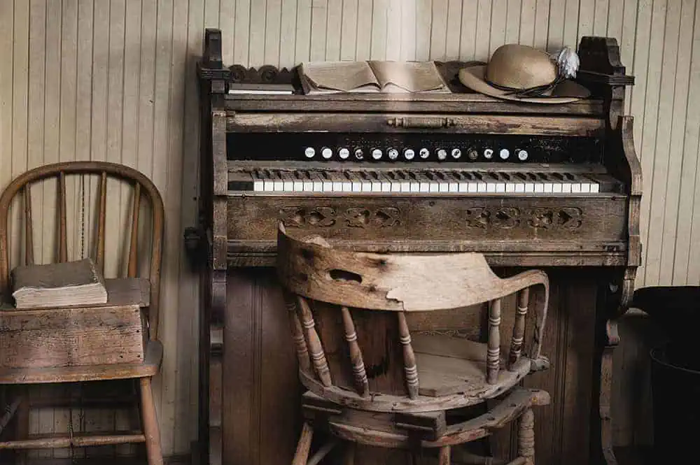 Antique pump organ with wooden frame, sheet music, and a straw hat on top beside rustic chairs.